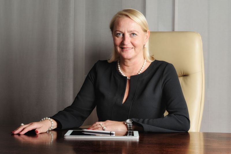 Frau mit blonden Haaren sitzt am Schreibtisch und lächelt, trägt ein schwarzes Oberteil.